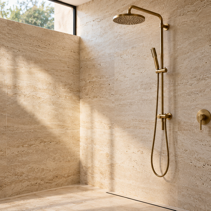 Brushed brass twin shower with an overhead rain head and handheld rail shower in a sunlit travertine bathroom.